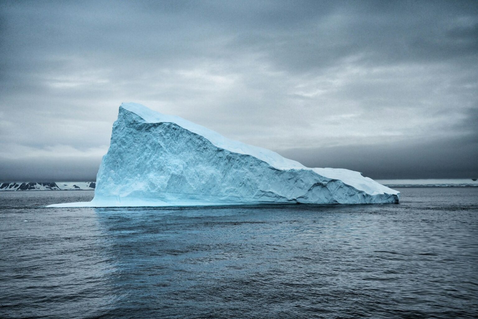 Iceberg Alley scenery beneath an overcast sky A pale blue iceberg floating in dark water beneath a cloudy Newfoundland sky